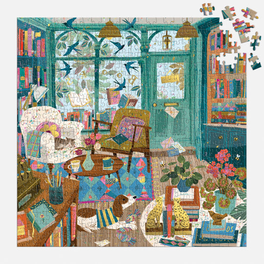 The Victoria Ball Bookshop Afternoon 500 Piece Foil Puzzle features a cozy, book-filled living room with big windows, plants, comfy chairs, a dog on a rug, and birds outside. Several pieces await placement in the top right corner.