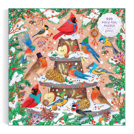 The Festive Feathers 500 Piece Foil Puzzle by Olga Akbarova showcases cardinals and robins on a snowy bird feeder, surrounded by pine branches, berries, pinecones, and apples on a pink backdrop.