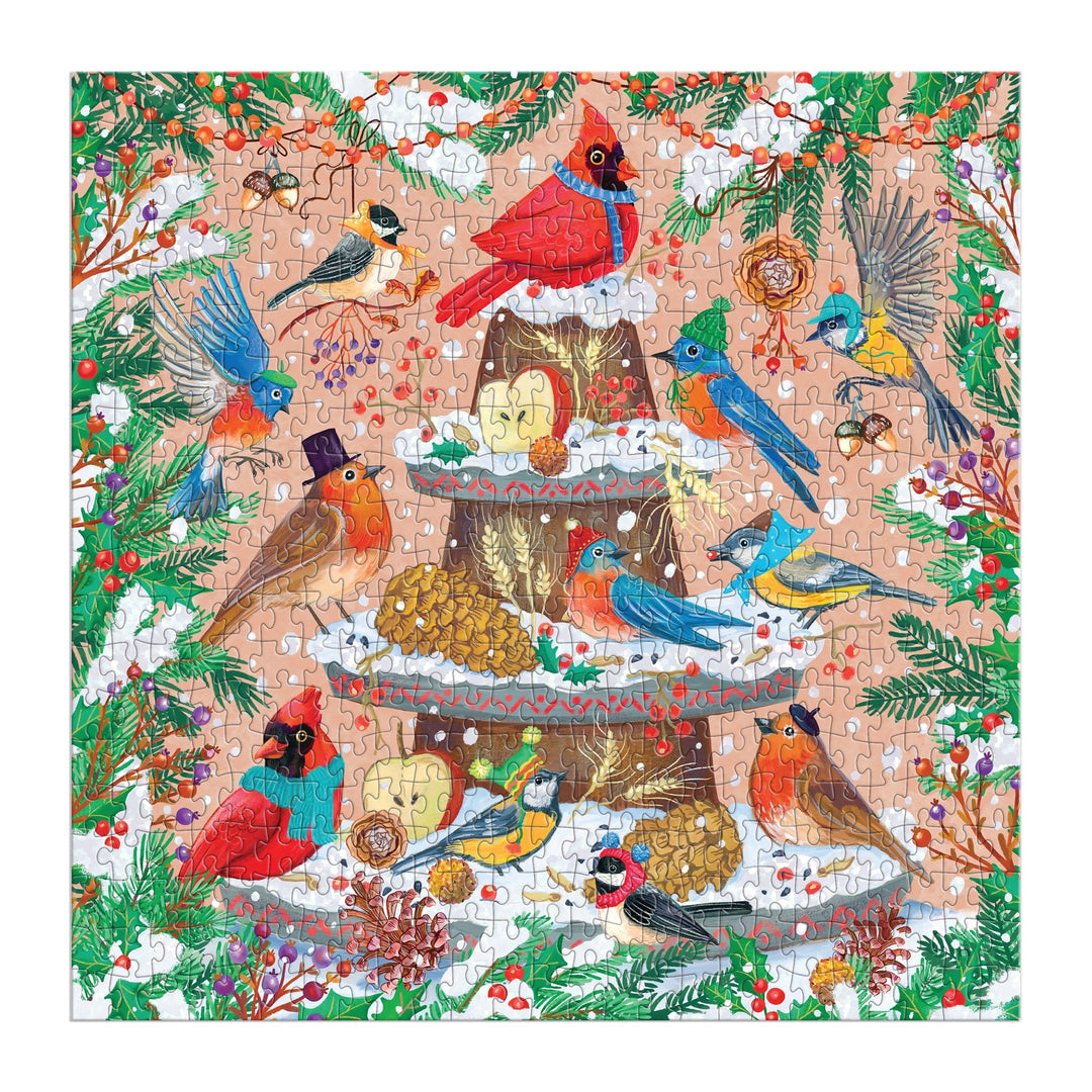 The Festive Feathers 500 Piece Foil Puzzle by Olga Akbarova features charming illustrations of cardinals, robins, and bluebirds gathered around a snowy tree stump with birdseed, pinecones, and festive winter decorations.