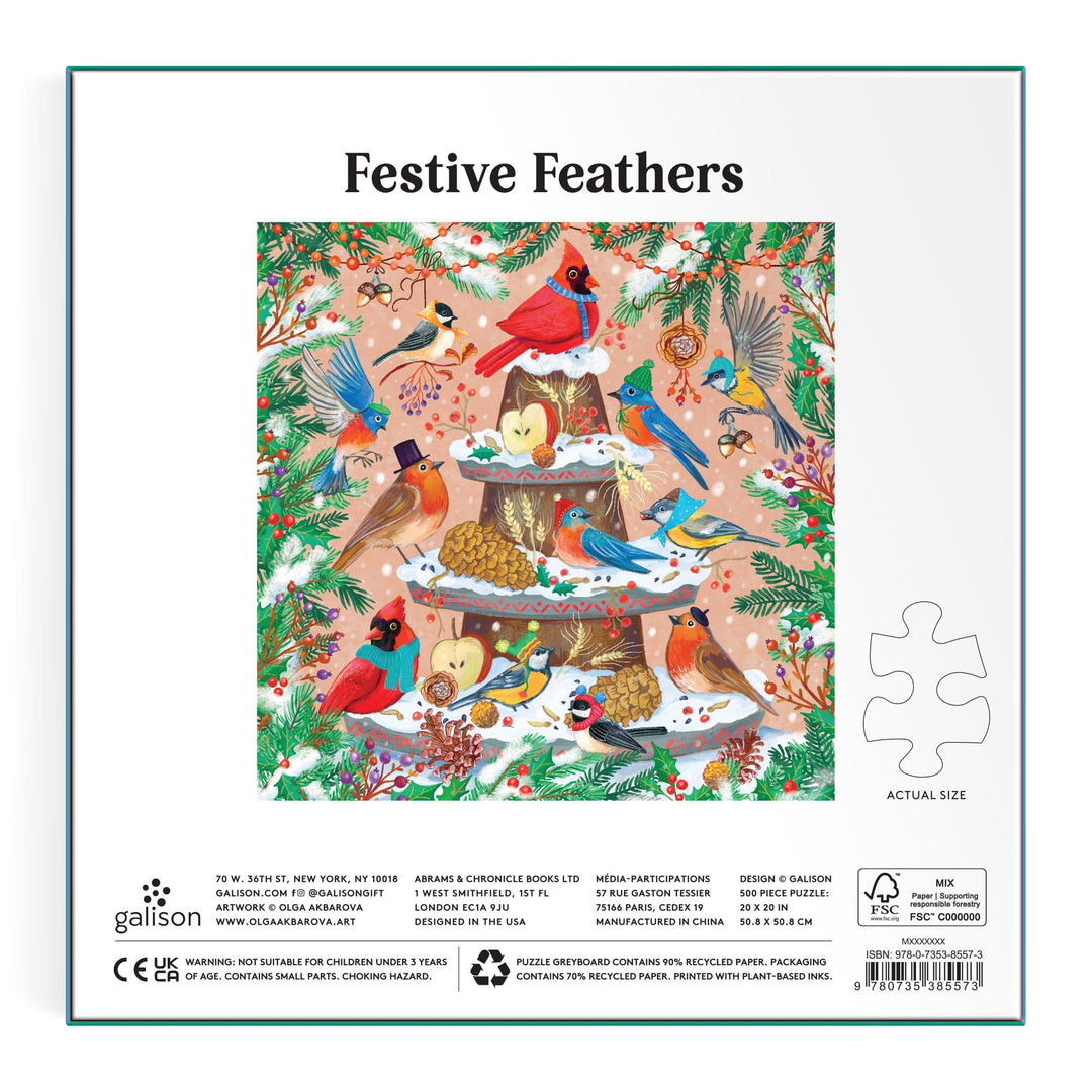 Festive Feathers 500 Piece Foil Puzzle by Olga Akbarova features cardinals and blue jays on a tiered feeder amid greenery, berries, and pine cones with metallic foil details, all set against a light pink background. Box and puzzle piece shown.