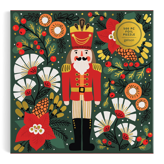The Merry Nutcracker 500 Piece Foil Puzzle by Sandra Hutter comes in a collector-quality box featuring festive artwork with flowers, pinecones, berries, and greenery. A gold sticker highlights the foil details.
