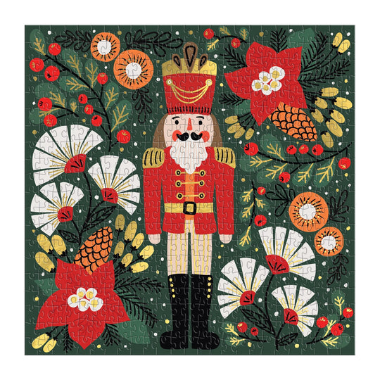 The Merry Nutcracker 500 Piece Foil Puzzle by Sandra Hutter features a nutcracker in a red uniform surrounded by festive flowers, pinecones, berries, foliage, and snowflakes—an ideal holiday-themed collector’s puzzle.