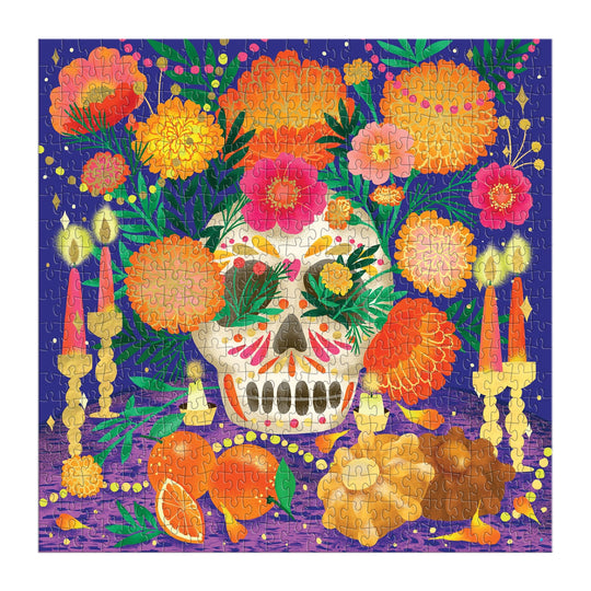 The Marisol Ortega Ofrenda del Día de Muertos 500 Piece Foil Puzzle features a vibrant purple background with a sugar skull, marigolds, candles, orange slices, and traditional offerings to celebrate Mexican heritage.