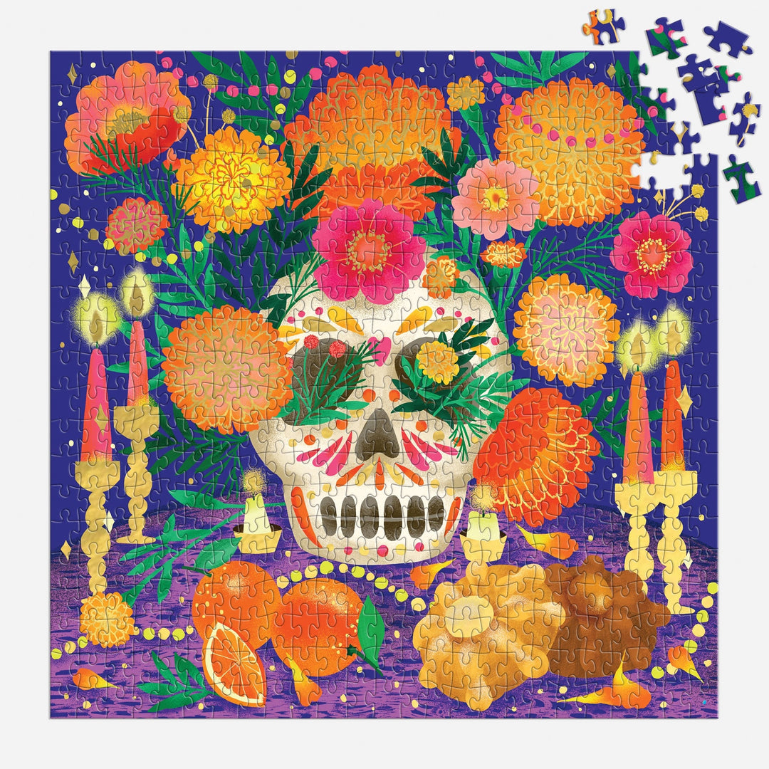 The Marisol Ortega Ofrenda del Día de Muertos 500 Piece Foil Puzzle features a sugar skull, marigolds, candles, orange slices, and bread on purple with metallic foil accents and some unfinished pieces in the top right corner.