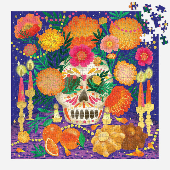 The Marisol Ortega Ofrenda del Día de Muertos 500 Piece Foil Puzzle features a sugar skull, marigolds, candles, orange slices, and bread on purple with metallic foil accents and some unfinished pieces in the top right corner.