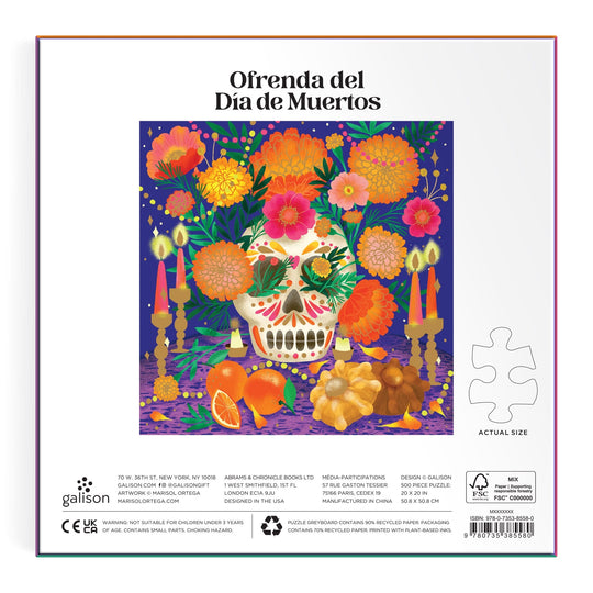 The Ofrenda del D’a de Muertos 500 Piece Foil Puzzle by Marisol Ortega features an illustrated altar with a decorated skull, candles, marigolds, fruit, leaves, and Spanish text on the box against a purple confetti background.