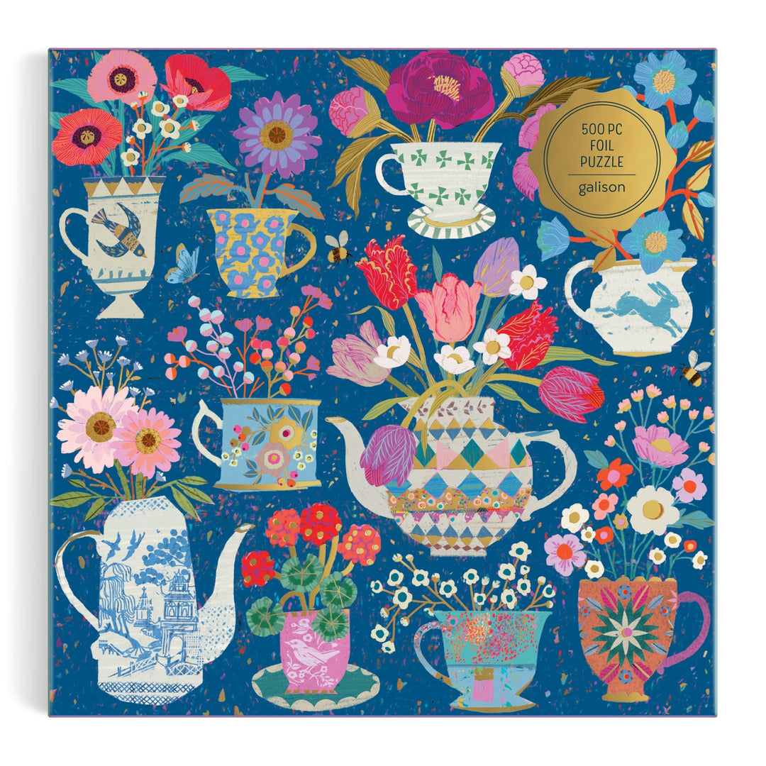 The Victoria Ball Potted Teacups 500 Piece Foil Puzzle features illustrated teapots, cups, and vases with colorful flowers on a blue background. A gold label highlights it as a foil puzzle by Victoria Ball.