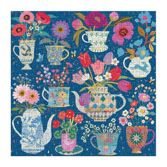 The Victoria Ball Potted Teacups 500 Piece Foil Puzzle by Victoria Ball features vibrant flowers in teapots, mugs, and pitchers on a blue background, all illustrated in colorful patterns and shades of pink, red, purple, and blue.
