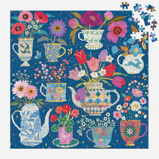 The Victoria Ball Potted Teacups 500 Piece Foil Puzzle features teapots, mugs, and pitchers brimming with vivid flowers on a deep blue background, with a few pieces yet to be placed in the upper right corner.