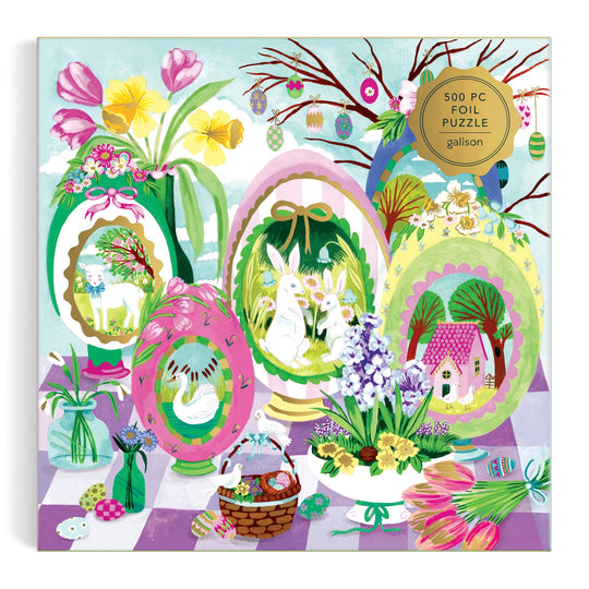 The Windows to Spring 500 Foil Piece Puzzle by Joanna Clay features large Easter eggs with animals and a cottage, surrounded by spring flowers, painted eggs, and treats. “500 pc foil puzzle” appears in the top right corner of the colorful box.