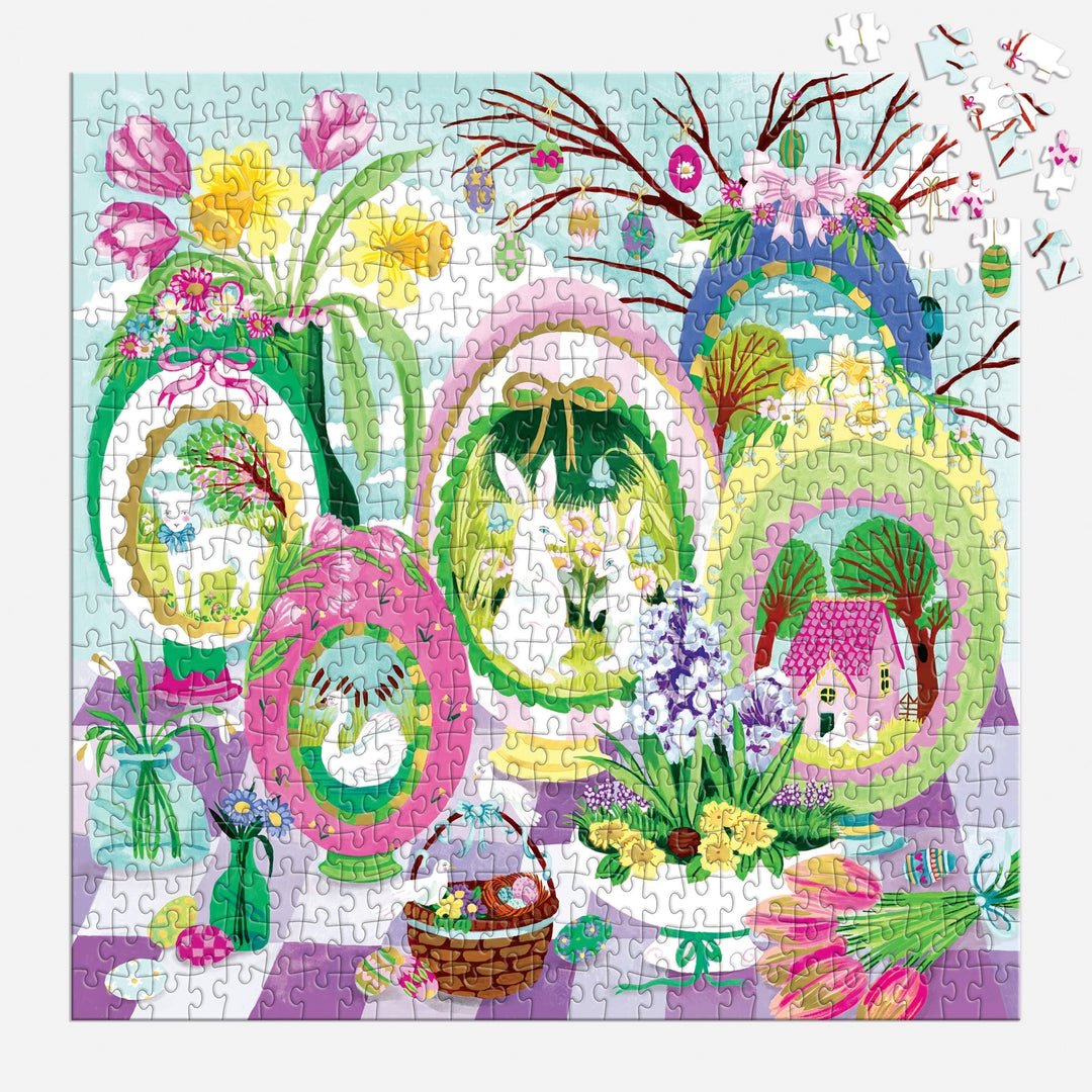 The Joanna Clay "Windows to Spring" 500 Foil Piece Puzzle features a vibrant spring scene with decorated egg-shaped frames, bunnies, flowers, and a house. Tulips and a basket adorn the foreground; a few pieces are missing in the top right corner.