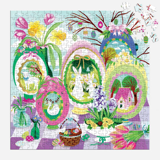 The Joanna Clay "Windows to Spring" 500 Foil Piece Puzzle features a vibrant spring scene with decorated egg-shaped frames, bunnies, flowers, and a house. Tulips and a basket adorn the foreground; a few pieces are missing in the top right corner.
