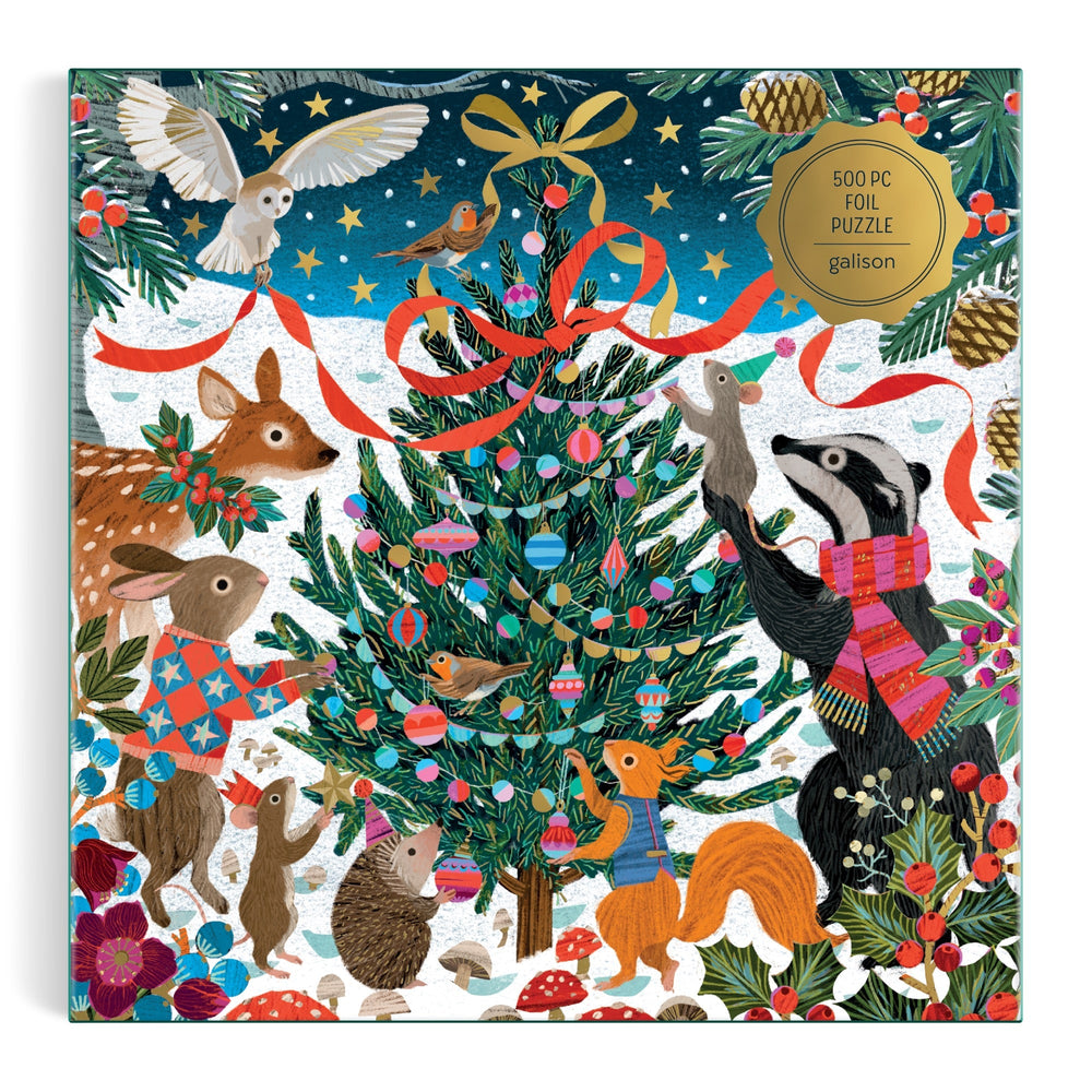 The Victoria Ball Woodland Merrymakers 500 Piece Foil Puzzle by Victoria Ball features woodland animals in scarves and sweaters decorating a Christmas tree with ornaments and ribbons in a snowy forest.