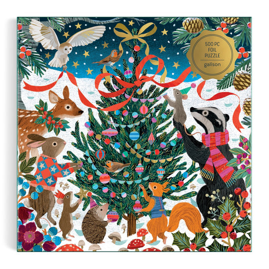 The Victoria Ball Woodland Merrymakers 500 Piece Foil Puzzle by Victoria Ball features woodland animals in scarves and sweaters decorating a Christmas tree with ornaments and ribbons in a snowy forest.