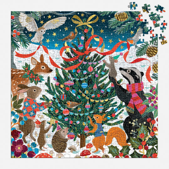 The Victoria Ball Woodland Merrymakers 500 Piece Foil Puzzle features woodland animals decorating a Christmas tree with ornaments and ribbons in a snowy forest, under festive plants and lit stars at night.