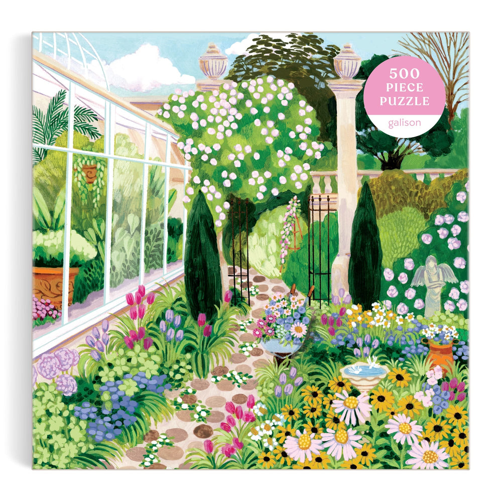 The Evergreen Gardens 500 Piece Puzzle by Joanna Clay features an illustration of a lush garden with colorful flowers, bushes, a stone path, fountain, and greenhouse on the box.