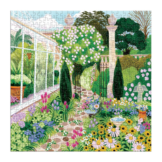The Joanna Clay Evergreen Gardens 500 Piece Puzzle features a vibrant garden scene with colorful flowers, green trees, a stone pathway, a white statue, birdbath, and a glass greenhouse beneath a bright blue sky.