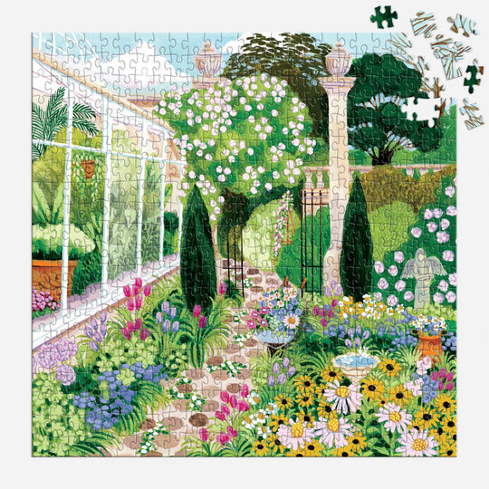 The Evergreen Gardens 500 Piece Puzzle by Joanna Clay depicts a vibrant garden path with blooming flowers, greenery, a statue, and a fountain near a glass conservatory; several pieces remain unfinished in the top right corner.