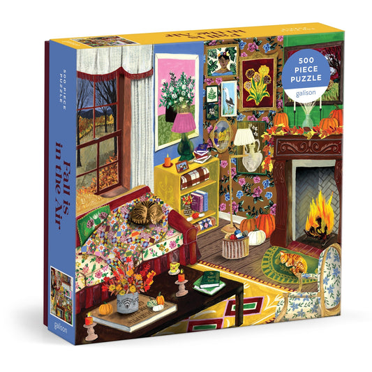 The Abbey Jacobs Fall is in the Air 500 Piece Puzzle features a cozy living room with a fireplace, cats, autumn decor, plants, books, and a floral sofa by a window with fall trees.