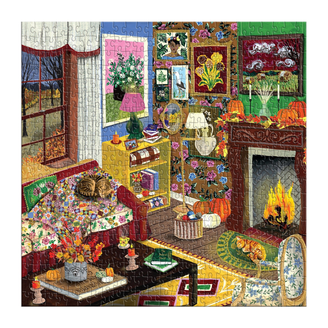 The "Fall is in the Air 500 Piece Puzzle" by Abbey Jacobs features a vibrant living room with a patterned couch, cozy pets, a glowing fireplace, floral accents, bookshelves, pumpkins, and large windows revealing an autumn scene.