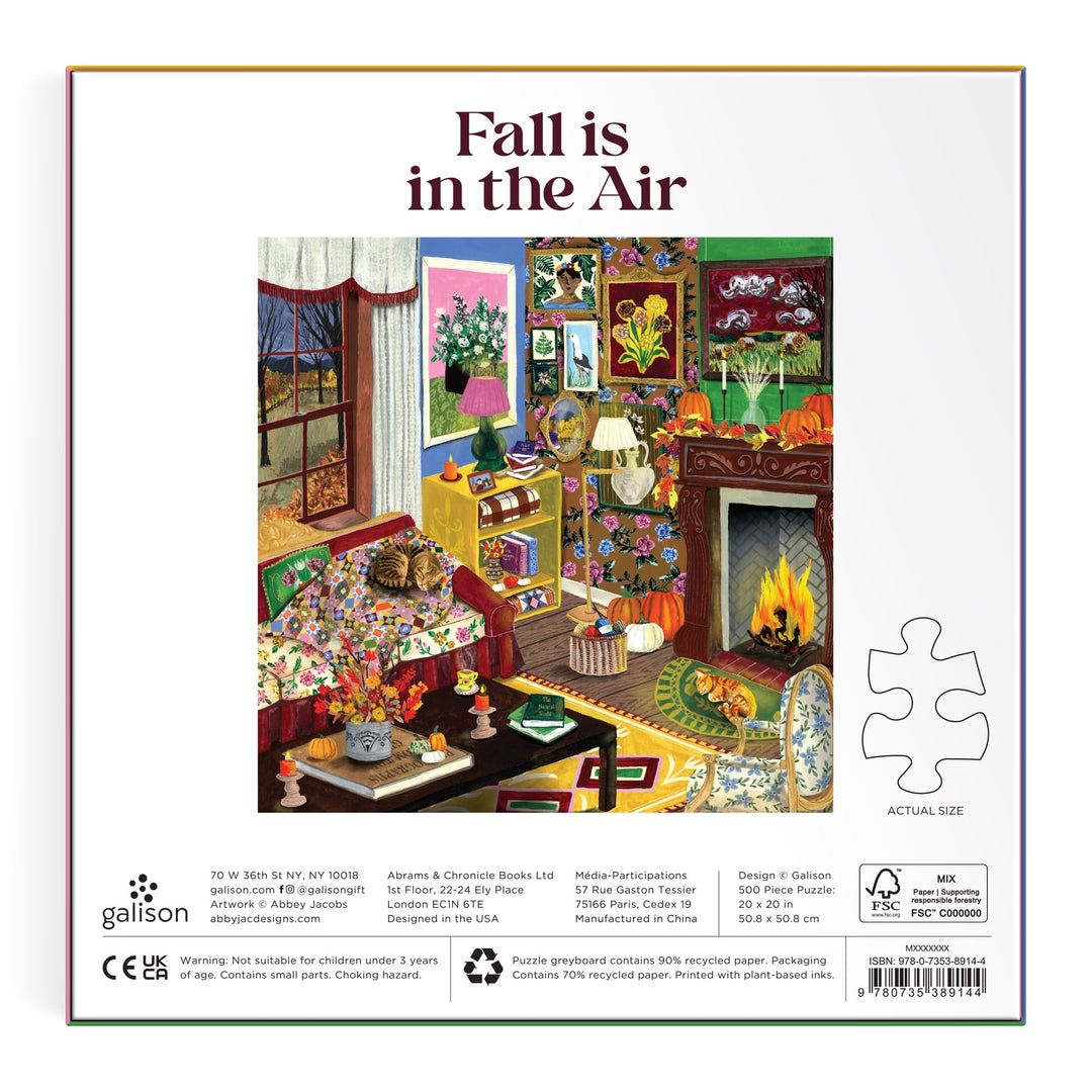 Enjoy assembling the "Fall is in the Air" 500 Piece Puzzle by Abbey Jacobs, featuring a cozy living room with a lit fireplace, armchair, tea table, autumn decor, and a view of vibrant red and orange foliage through the window.