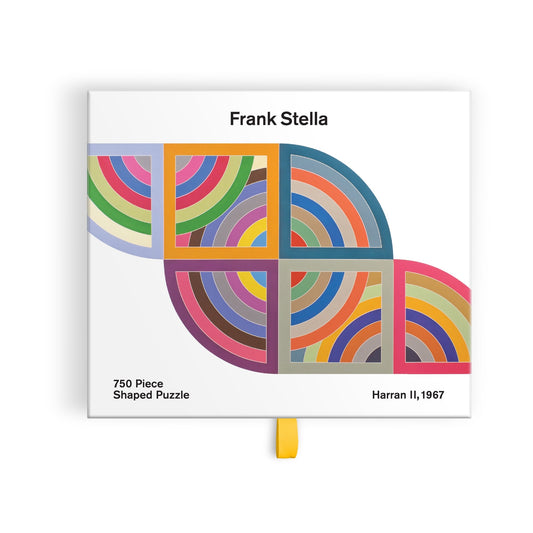 The Frank Stella Harran II 750 Piece Shaped Puzzle in Drawer Box features bold, rainbow-colored arcs from Stella’s geometric artwork Harran II, 1967. Box text: “Frank Stella,” “750 Piece Shaped Puzzle,” and “Harran II, 1967.”.