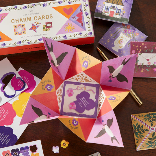A colorful origami-style paper fortune teller with bird illustrations sits among refined stationery, decorative charm cards from Joy Laforme, envelopes, paper flowers, and a pen. The Joy Laforme Charm Cards box is also in view.
