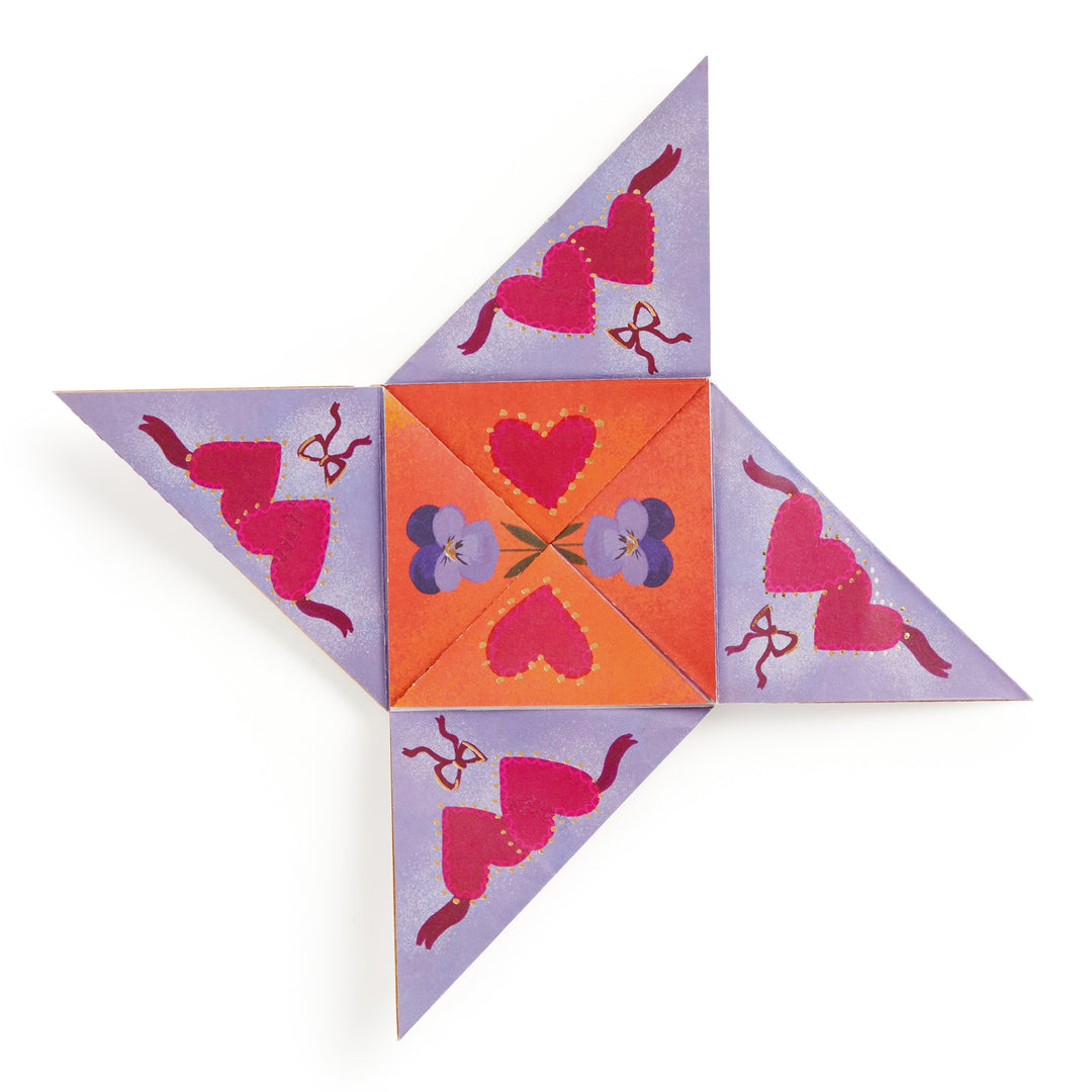 A vibrant paper fortune teller inspired by Joy Laforme Charm Cards features red hearts, purple flowers, and bows on a purple-orange background, unfolded to reveal its triangular flaps. Brand: Joy Laforme.