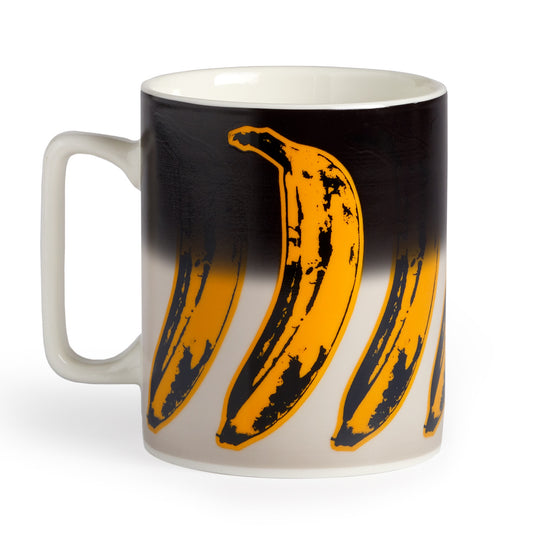 The Andy Warhol Banana Color Changing Ceramic Mug by Andy Warhol features a wraparound pop art design of large yellow bananas with black outlines on a gradient black-to-white background.