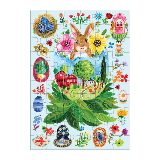 The Artisanal Eggs Greeting Card Puzzle by Jennifer O Lewis / August Wren features a rabbit, flowers, birds, and festive eggs with a village of red houses and green trees, celebrating spring and Easter wishes.