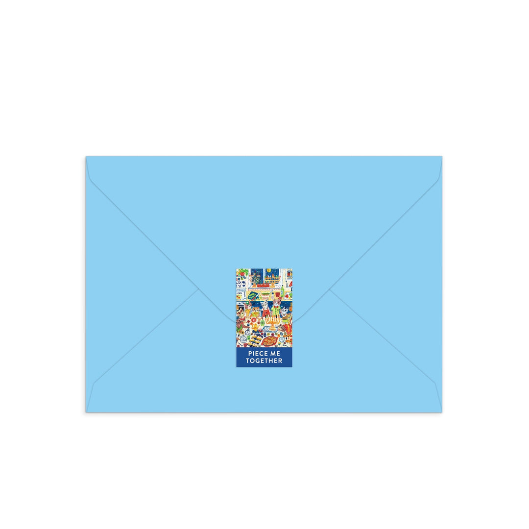 A closed light blue envelope with a festive feline sticker and the text PIECE ME TOGETHER holds the Happy Hanukcats Greeting Card Puzzle by Anna Ivanir—an interactive Hanukkah card with a playful cat-themed puzzle inside.