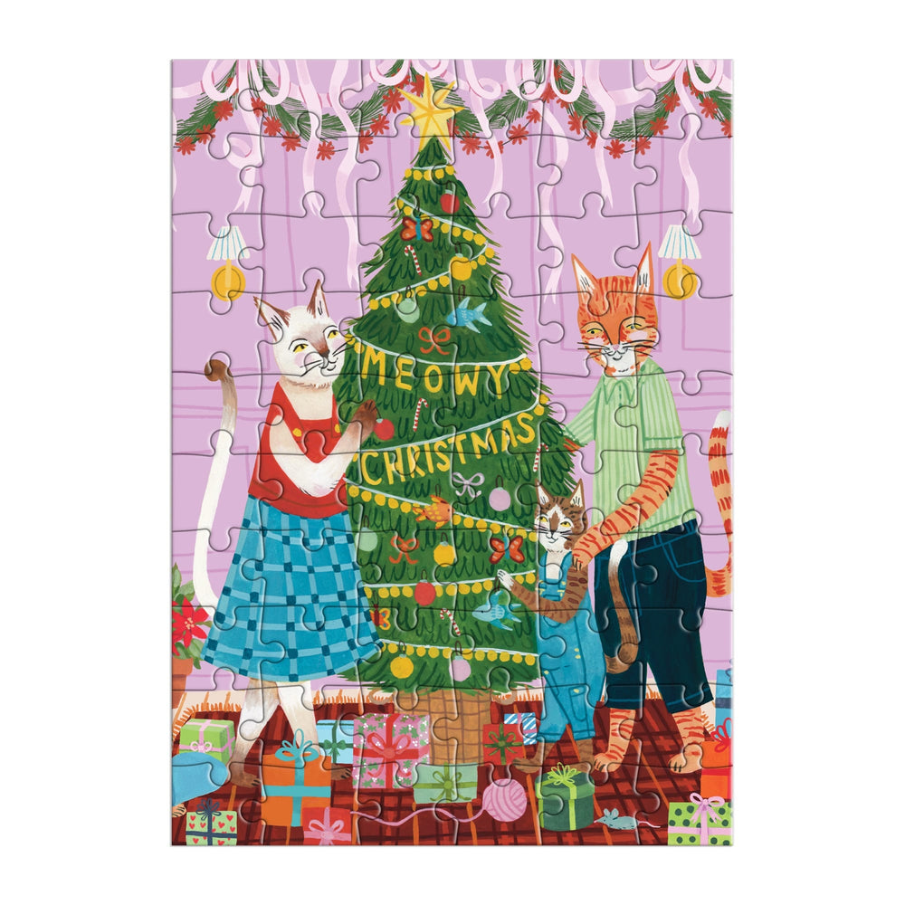 The Meowy Christmas Greeting Card Puzzle by Joanna Clay features three cats—two adults and a kitten—decorating a Christmas tree with festive décor, wrapped gifts, and a Meowy Christmas banner on a cheerful pink background.