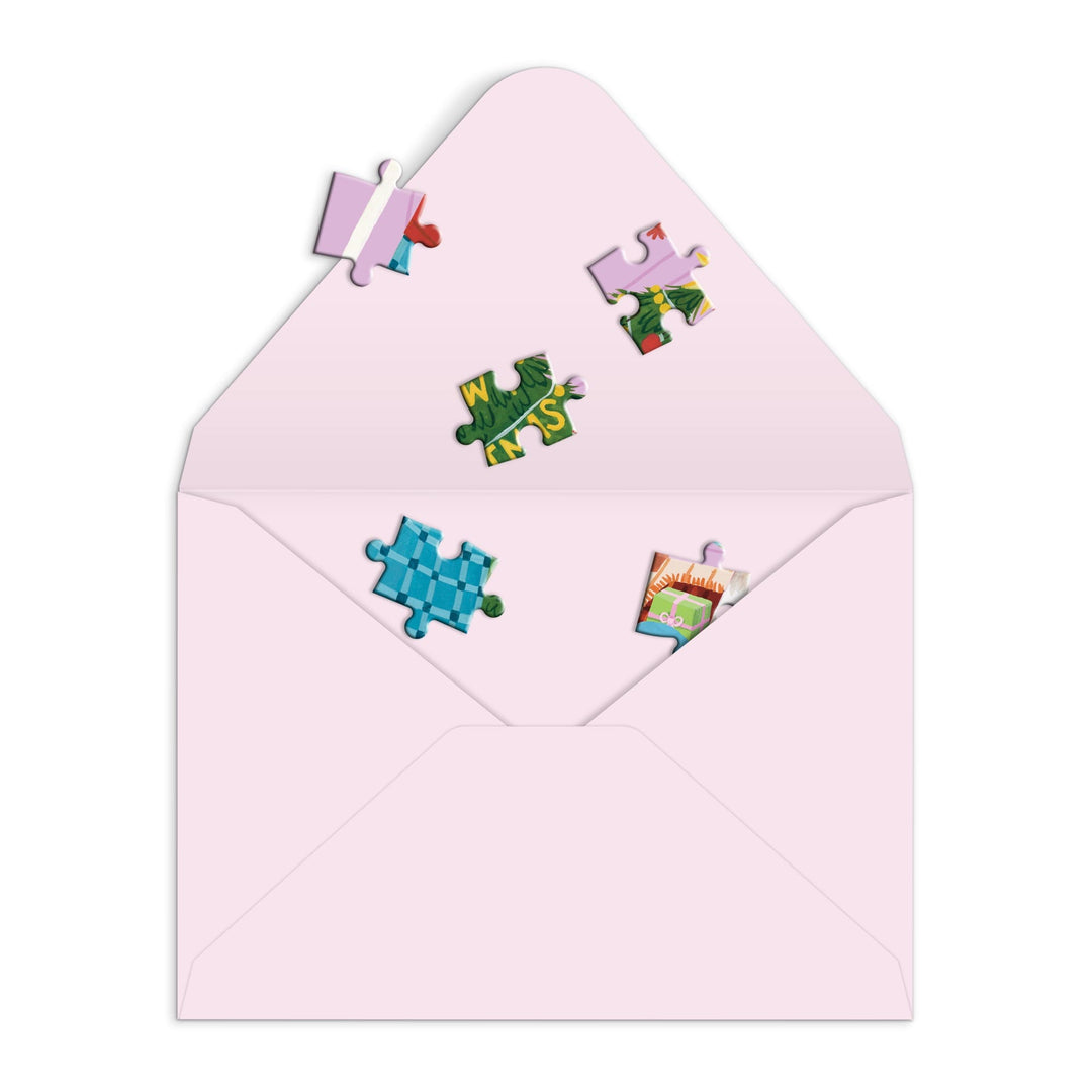An open pink envelope with six colorful puzzle pieces from Joanna Clay’s Meowy Christmas Greeting Card Puzzle, scattered inside and on the flap against a white background—ideal for fans of interactive entertainment.