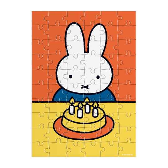 The Miffy Birthday Greeting Card Puzzle by Miffy features a white cartoon bunny with upright ears at a table, celebrating with a yellow cake topped with five lit candles against an orange and yellow background.