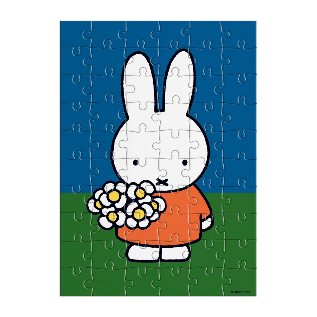 The Miffy with Flowers Greeting Card Puzzle by Miffy features a white cartoon rabbit in an orange dress, holding white flowers on green grass with a blue sky background.