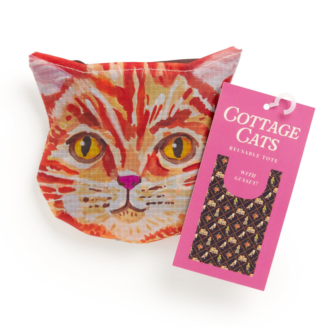 A pouch shaped like an orange tabby cat’s face sits beside a pink tag that reads "Cottage Cats Packable Nylon Tote" by Anja Riebensahm, featuring a cat-face pattern on the bag.
