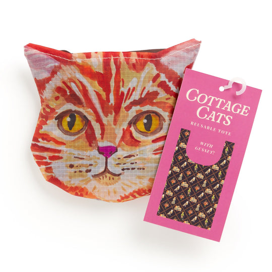 A pouch shaped like an orange tabby cat’s face sits beside a pink tag that reads "Cottage Cats Packable Nylon Tote" by Anja Riebensahm, featuring a cat-face pattern on the bag.