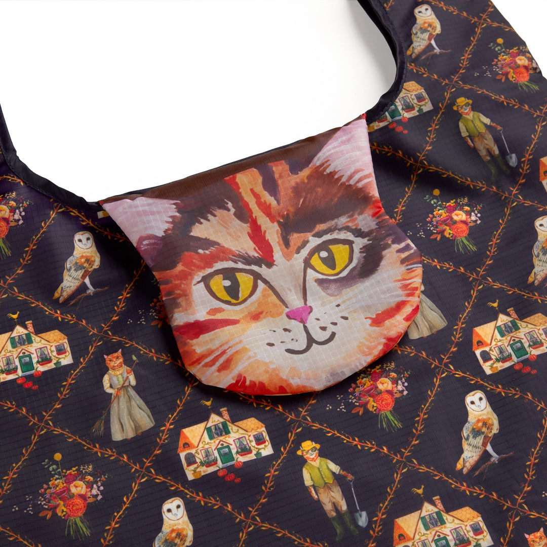 The Cottage Cats Packable Nylon Tote by Anja Riebensahm is a reusable bag with a flap featuring a realistic cat face and dark diamond-patterned fabric adorned with whimsical cats in clothes, owls, bouquets, and tiny houses.