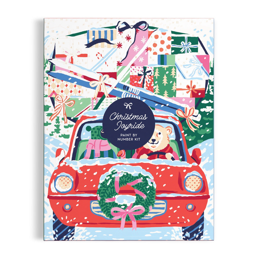 A festive illustration on the “Christmas Joyride 9 x 12" Paint by Number Kit” by Emily Taylor features a bear driving a red, gift-filled car with skis and trees, set in snowy scenery for holiday fun.