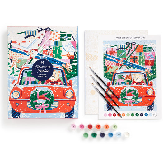 The Emily Taylor Christmas Joyride 9 x 12" Paint by Number Kit features a car with a wreath, two dogs, and gifts. Includes canvas, paint guide, brushes, and numbered paint pots in assorted colors.