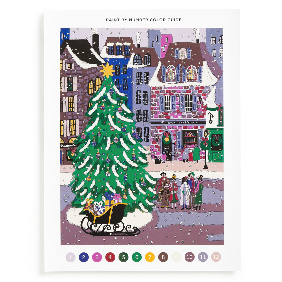 The Joy Laforme Christmas Square 9 x 12 Paint by Number Kit features a snowy town square with a decorated tree, sleigh of gifts, festive buildings, and people. Numbered color circles are displayed below the vibrant holiday scene.