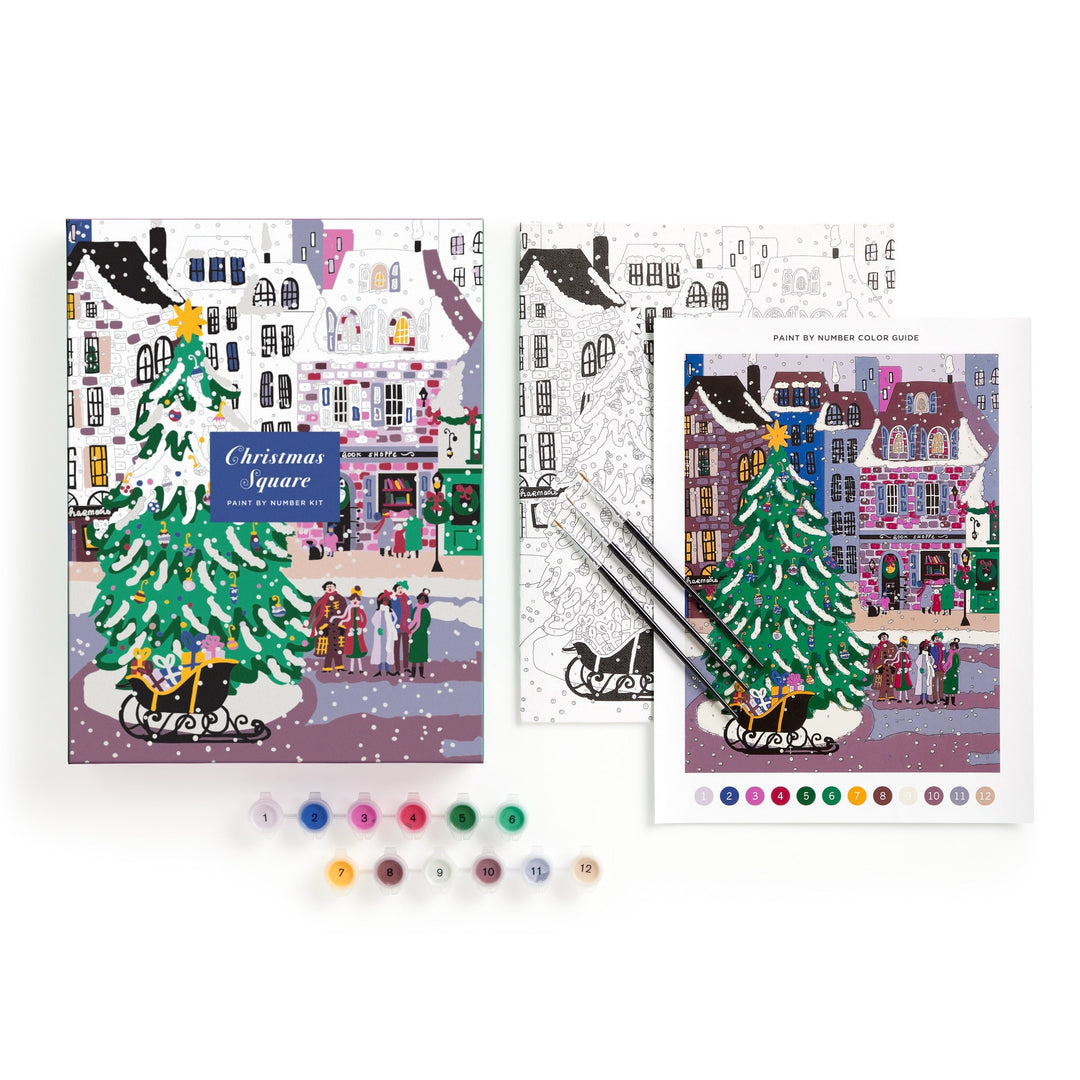 The Joy Laforme Christmas Square 9 x 12 Paint by Number Kit features a festive holiday street scene with a decorated tree, paint pots, brushes, and two illustrated sheets—one finished and one black-and-white—on a white background.