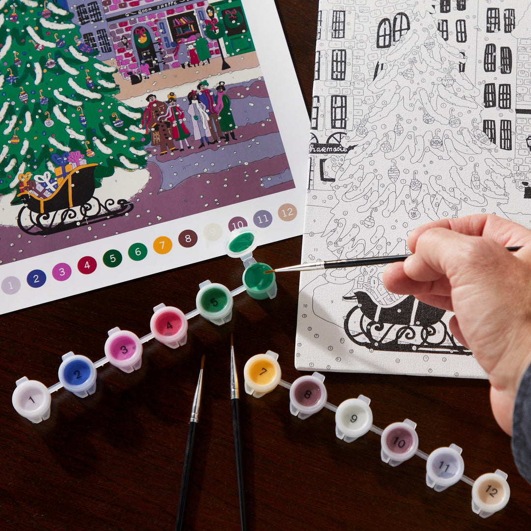 A hand holds a brush over the Joy Laforme Christmas Square 9 x 12 Paint by Number Kit canvas with a festive tree scene. Nearby are paint pots, brushes, and a color reference—ideal for making your own holiday décor. Brand: Joy Laforme.