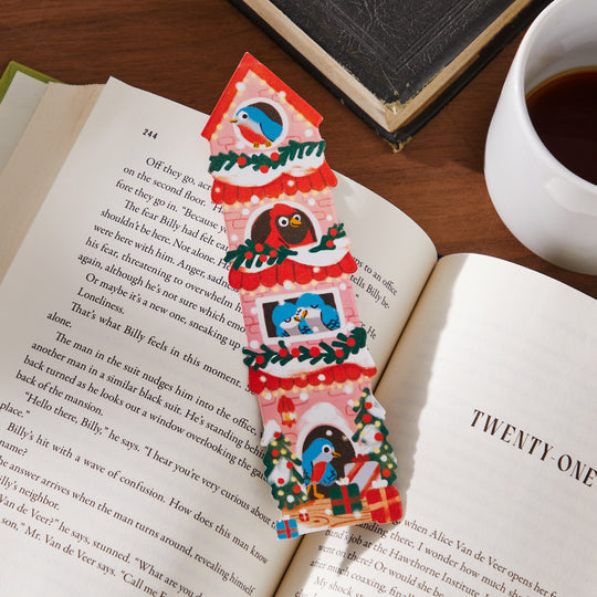 The Éléna Comte Seasons Tweetings Paper Bookmark—festively adorned with birds and holiday décor—rests on an open book beside another book and a cup of black coffee, making it a charming holiday reading accessory.