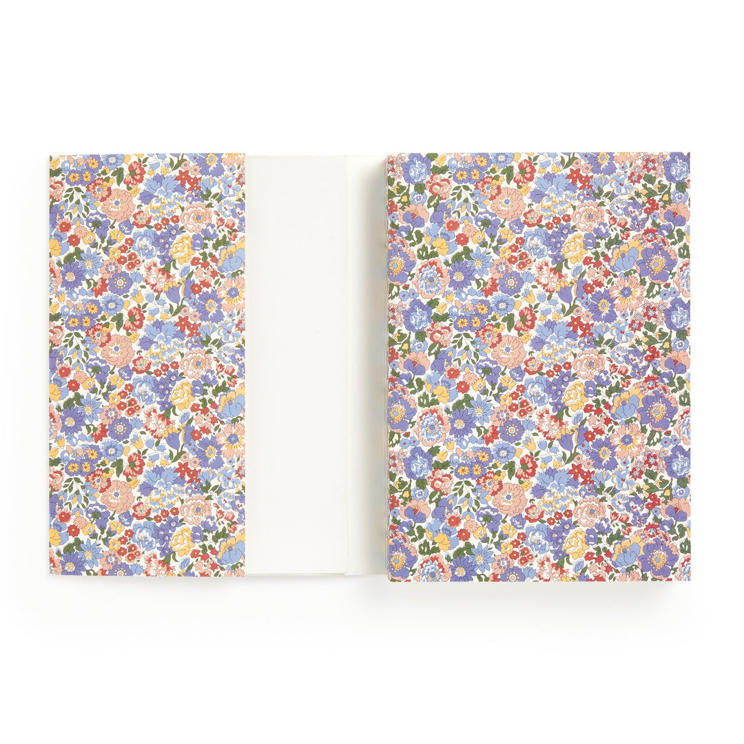 The Liberty London Nell, Annie and May Premium A5 Notebook is open, revealing a colorful floral-patterned cover and endpapers with small pink, blue, red, and orange flowers. Inside are blank white pages.