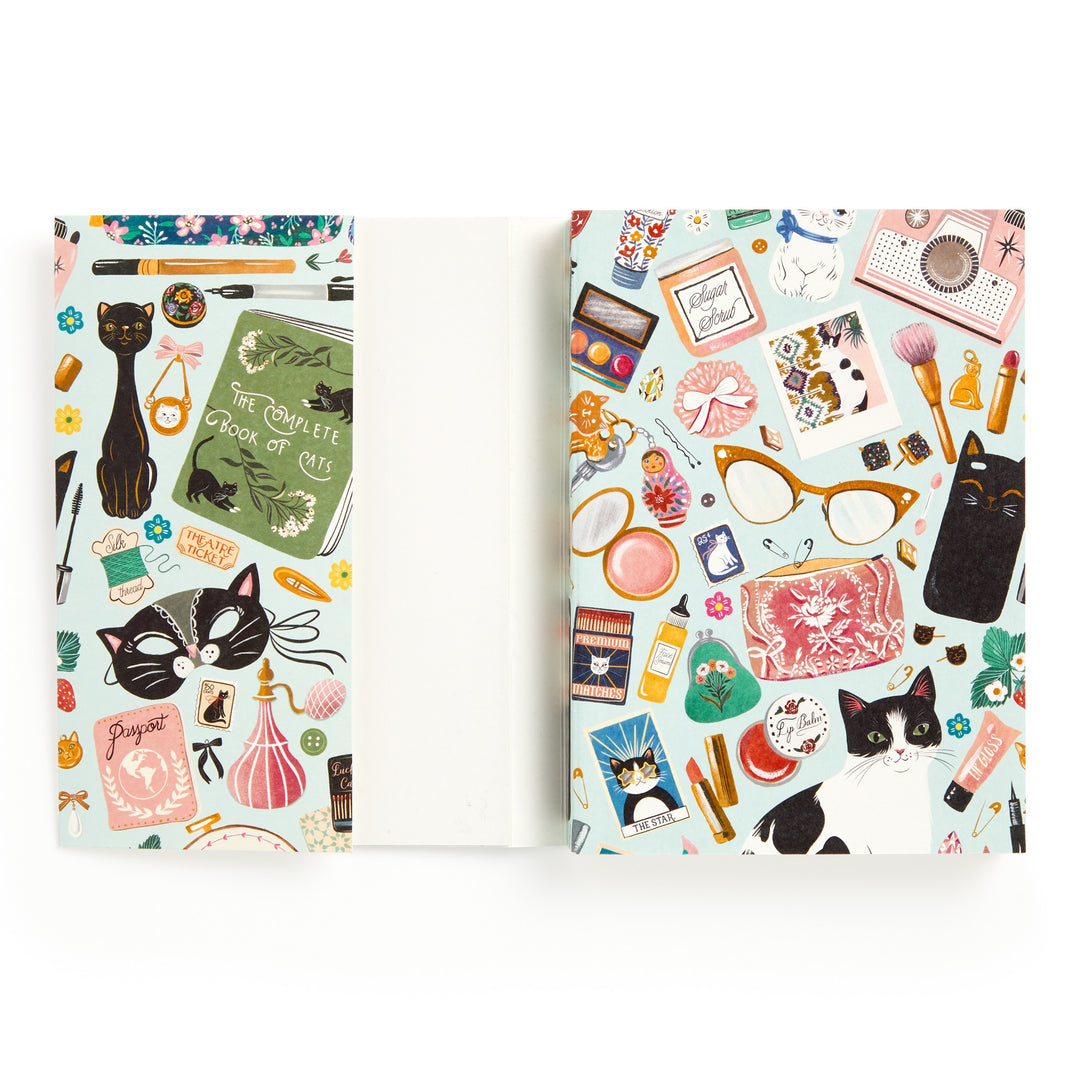 The Christine De Carvalho Purrfect Accessories Premium A5 Notebook features a vibrant illustrated cover with cats and cat-themed items—glasses, makeup, cameras, books, and stationery—on a white background.