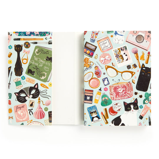 The Christine De Carvalho Purrfect Accessories Premium A5 Notebook features a vibrant illustrated cover with cats and cat-themed items—glasses, makeup, cameras, books, and stationery—on a white background.