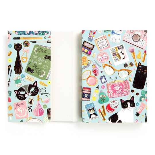 The Christine De Carvalho Purrfect Accessories Premium A5 Notebook features illustrated covers with colorful objects, makeup, books, and playful cats on a light blue background. The visible spine and pages add a stylish touch.