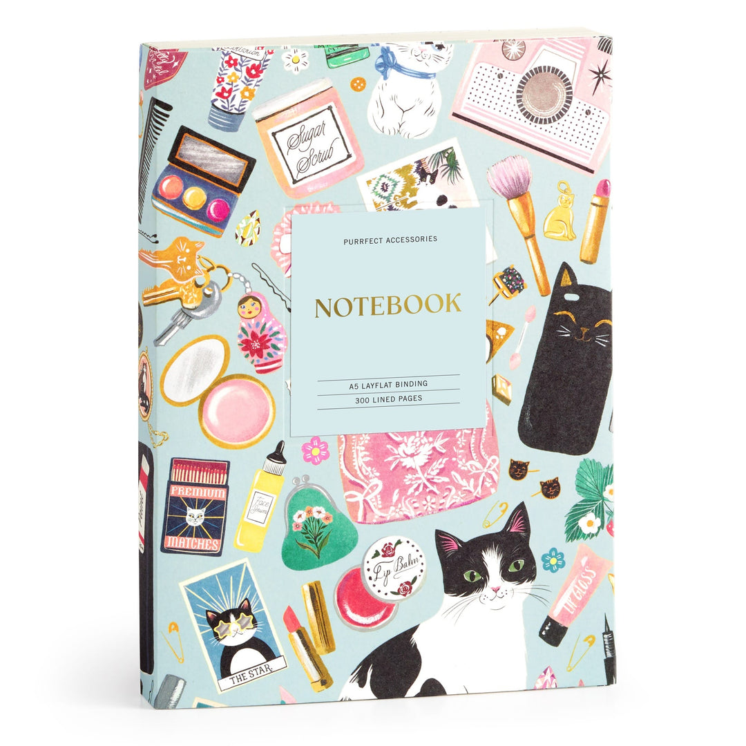 The Christine De Carvalho Purrfect Accessories Premium A5 Notebook features a colorful cover with cats, makeup, perfume bottles, and accessories on light blue, plus 300 lined premium pages.