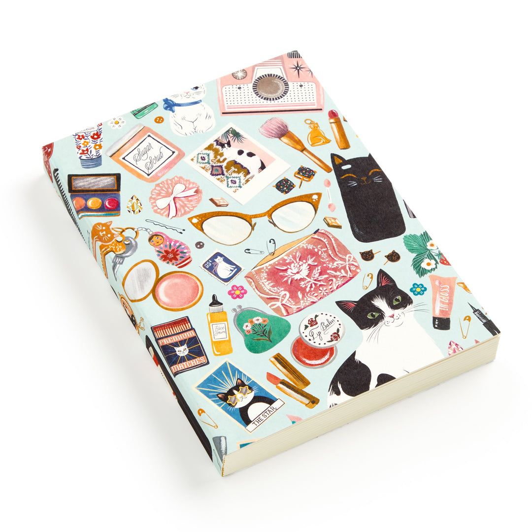 The Purrfect Accessories Premium A5 Notebook by Christine De Carvalho features a vibrant cover with chic feline illustrations, glasses, cosmetics, makeup items, perfume, brushes, and flowers on a light blue background.
